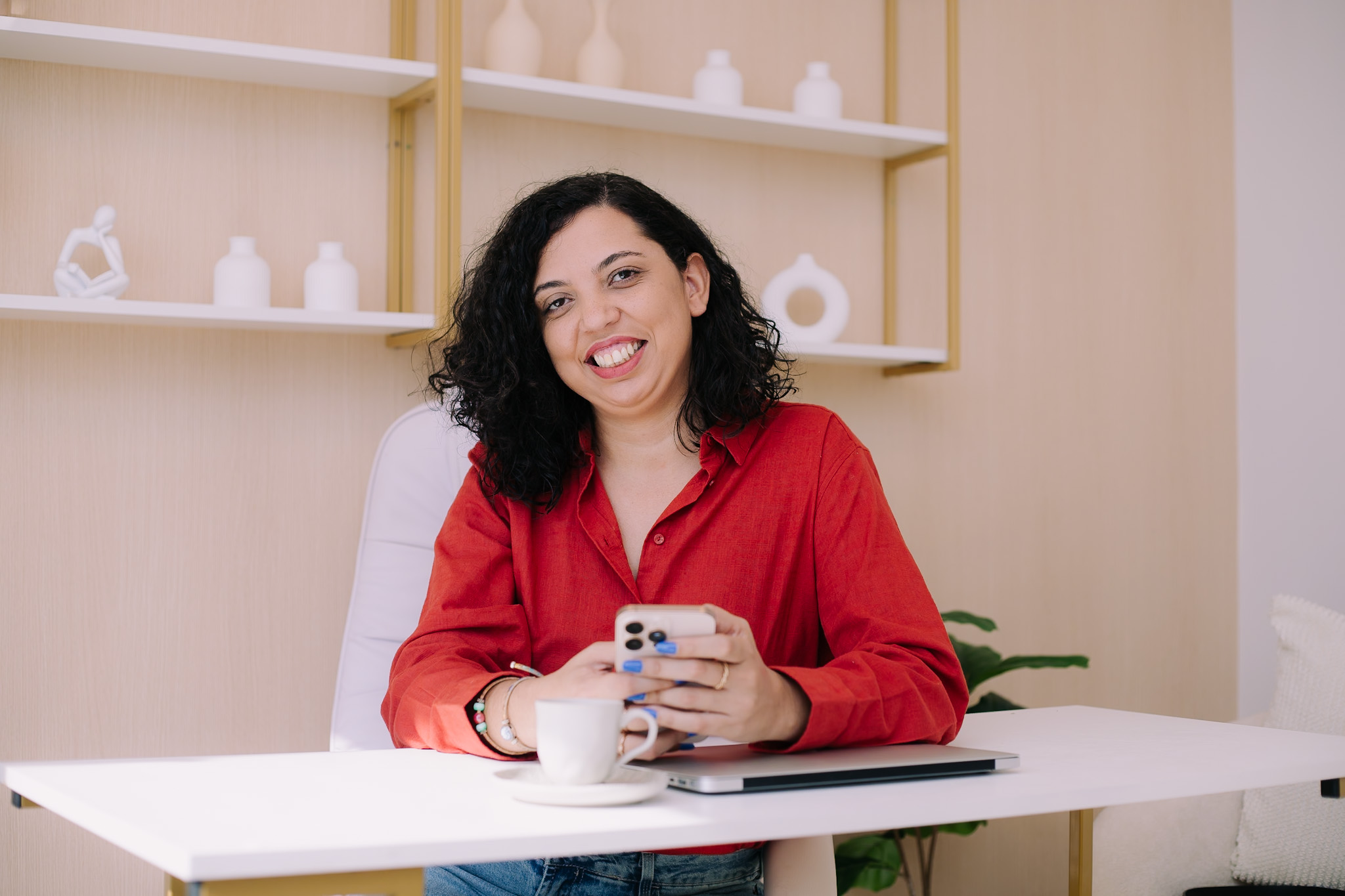 Nangela Lima sorrindo à mesa de trabalho, com uma xícara e o celular nas mãos